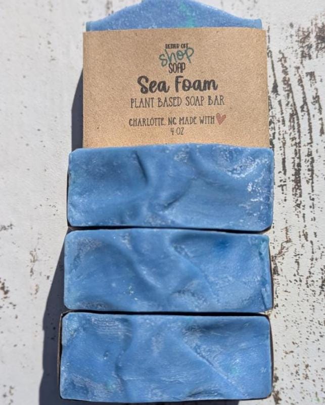 A stack of three Sea Foam soap bars from Better Off Soap Shop, showcasing the blue and teal tones with a marbled, slightly textured pattern on the top. The top bar is wrapped in eco-friendly kraft paper packaging, while the bottom two soaps reveal their swirled, glossy surface with subtle iridescent specks. Displayed on a rustic white wooden surface under bright natural lighting, casting soft shadows.