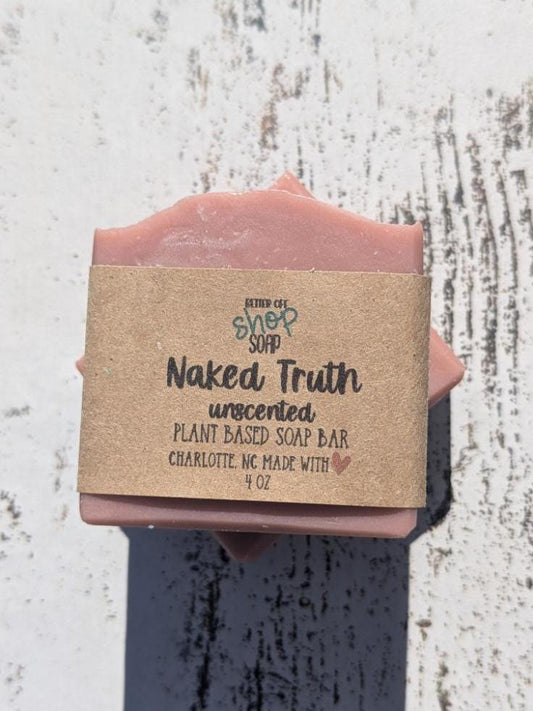Handcrafted, unscented soap bar from Better Off Soap Shop, labeled &#39;Naked Truth.&#39; This plant-based soap has a soft, muted pink hue and a slightly textured, wavy top. It is wrapped in eco-friendly brown kraft paper with black and teal lettering. The soap rests on a rustic, whitewashed wooden surface with natural light casting a gentle shadow. Made in Charlotte, NC, with natural ingredients, this 4 oz bar is perfect for those who prefer fragrance-free, skin-friendly cleansing.