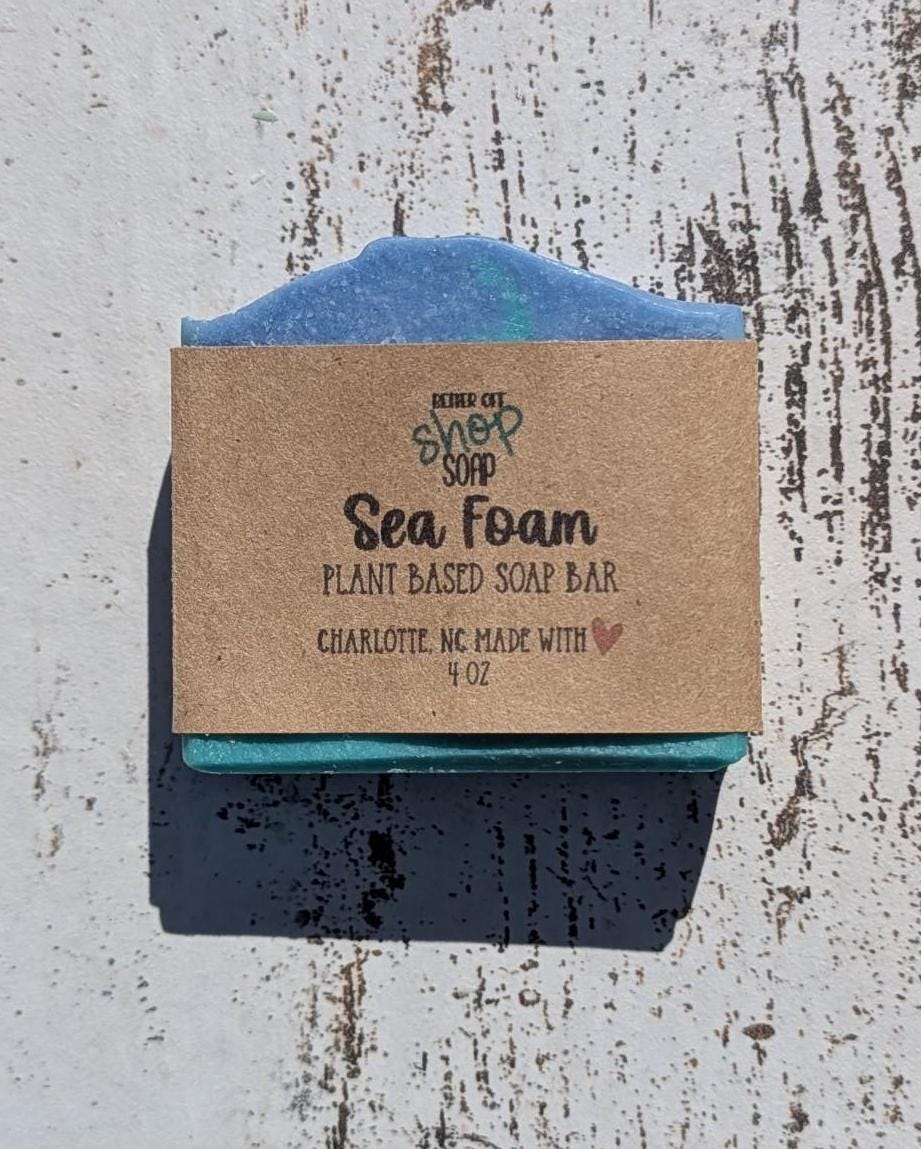 A handcrafted Sea Foam soap bar from Better Off Soap Shop, featuring a two-toned blue and green design with a soft, wavy top. The soap is wrapped in eco-friendly kraft paper packaging with black and green branding, highlighting it as a plant-based, handmade soap from Charlotte, NC. Photographed on a rustic white wooden surface under natural sunlight, casting a crisp shadow.