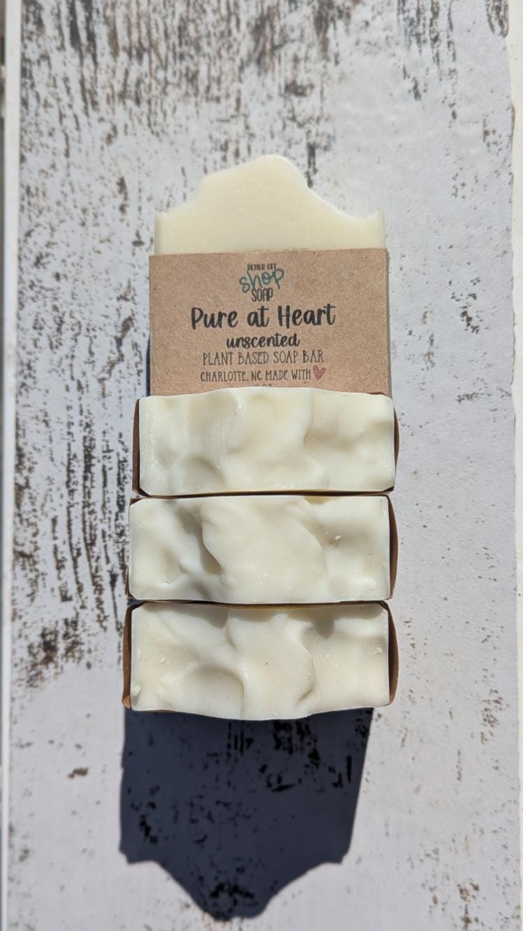 Close-up image of a handcrafted, unscented soap bar from Better Off Soap Shop, labeled &#39;Pure at Heart.&#39; This plant-based soap bar is wrapped in eco-friendly brown kraft paper with black and teal lettering. It is made in Charlotte, NC, with natural ingredients. The creamy white soap has a smooth, slightly wavy top, and is placed on a wooden background with other soap bars in the background. Perfect for those with sensitive skin or who prefer fragrance-free skincare.