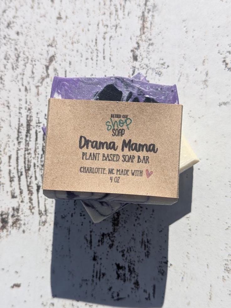 A handmade cold process soap bar in purple, black, and white swirls, standing upright on a white wooden background. Wrapped in an eco-friendly recyclable paper band.