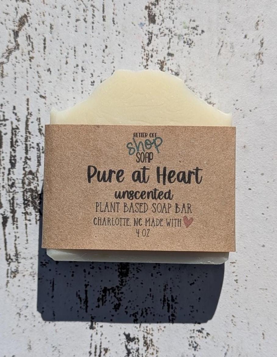 Close-up image of a handcrafted, unscented soap bar from Better Off Soap Shop, labeled &#39;Pure at Heart.&#39; This plant-based soap bar is wrapped in eco-friendly brown kraft paper with black and teal lettering. It is made in Charlotte, NC, with natural ingredients. The creamy white soap has a smooth, slightly wavy top, and is placed on a wooden background. Perfect for those with sensitive skin or who prefer fragrance-free skincare.