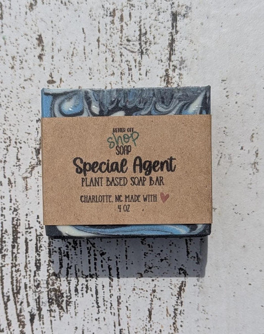 Special Agent handmade soap bar wrapped in a kraft paper label, featuring black, white, and blue swirls, placed on a white distressed wood background.