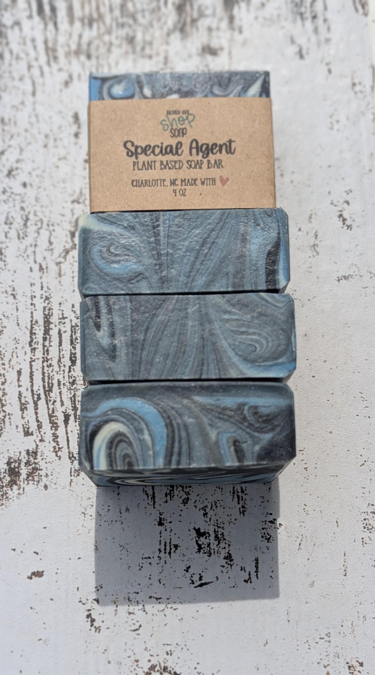 Special Agent handmade soap bar wrapped in a kraft paper label, featuring black, white, and blue swirls, placed on a white distressed wood background.